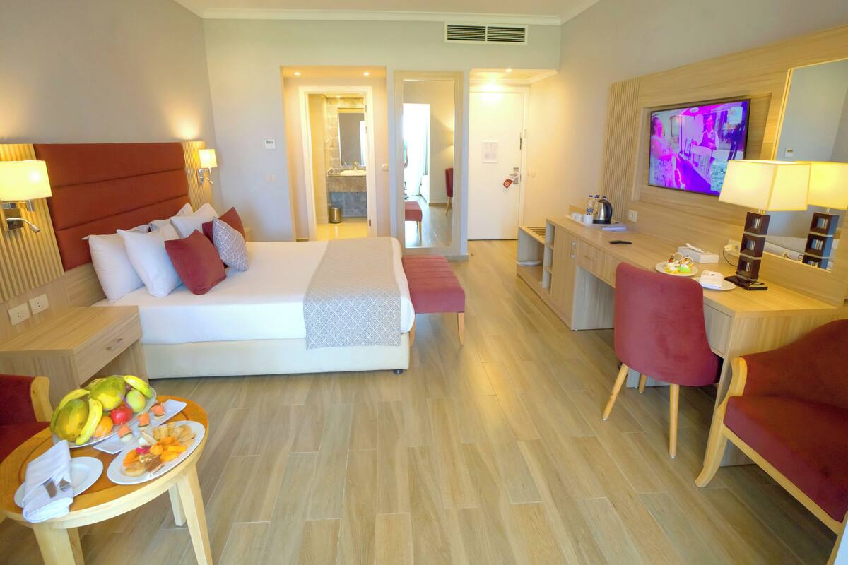A hotel room with a bed, a desk, a TV, and a dining table with fruit.