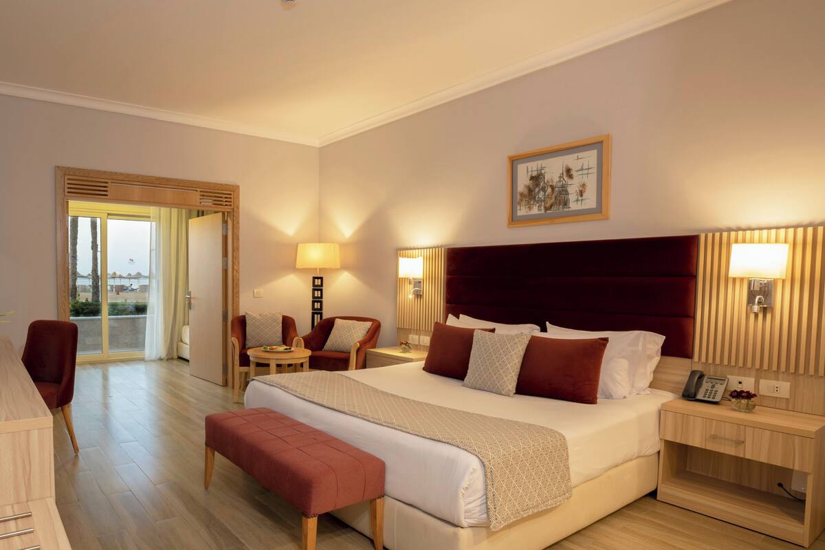 A hotel room with a large bed, a desk, a chair, a bench, and a view of the outside.