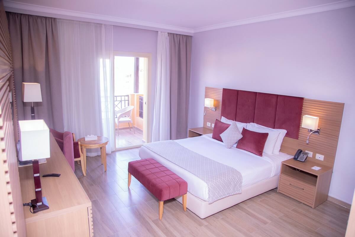 A hotel room with a large bed, a bedside table with a lamp, a small desk with a lamp, a chair, and a bench.