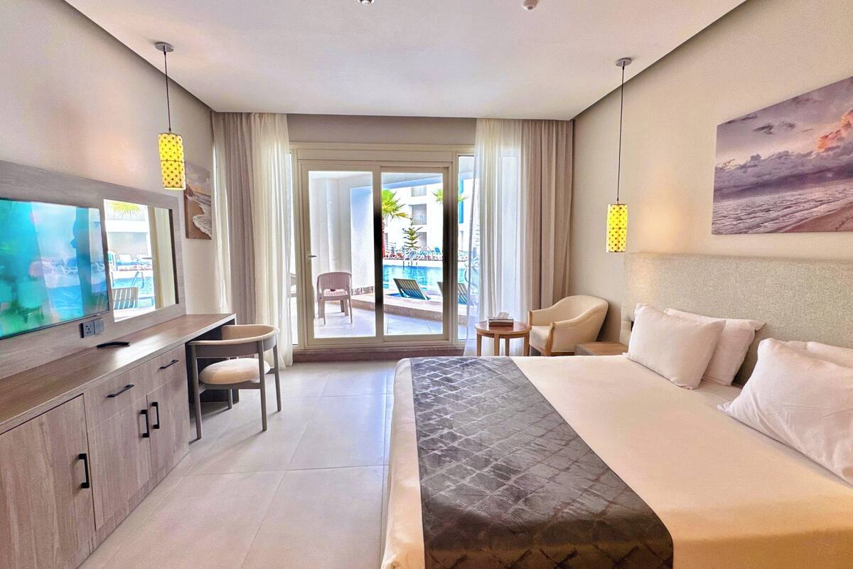 A hotel room with a bed, a TV, a desk, a chair, a small table, and a view of a pool and outdoor area.