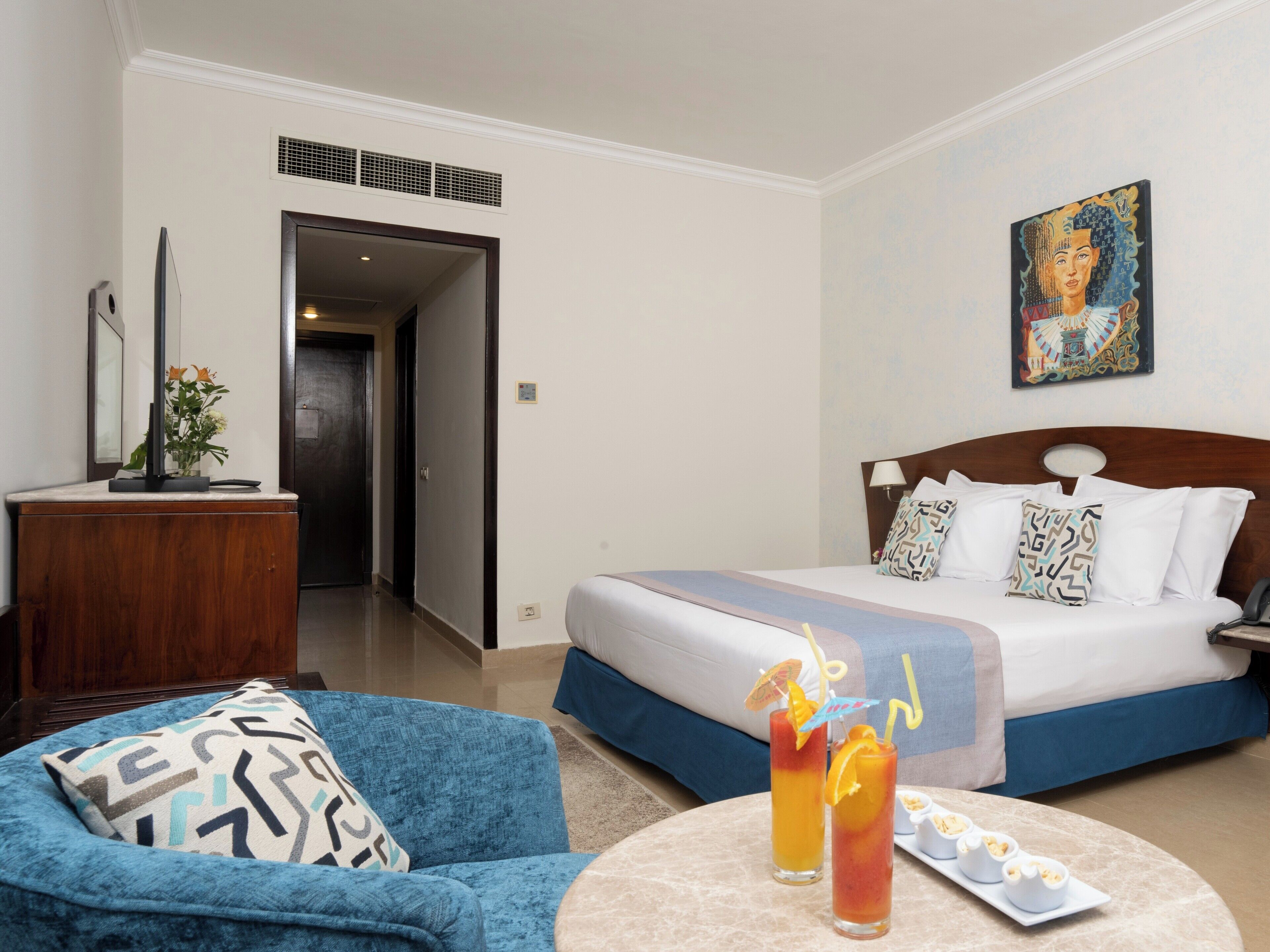 A hotel room with a bed, a desk, a chair, a painting, and a table with drinks and snacks.