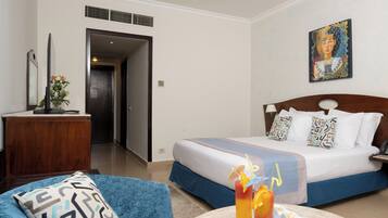 A hotel room with a bed, a desk, a chair, a painting, and a table with drinks and snacks.