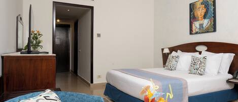 A hotel room with a bed, a desk, a chair, a painting, and a table with drinks and snacks.