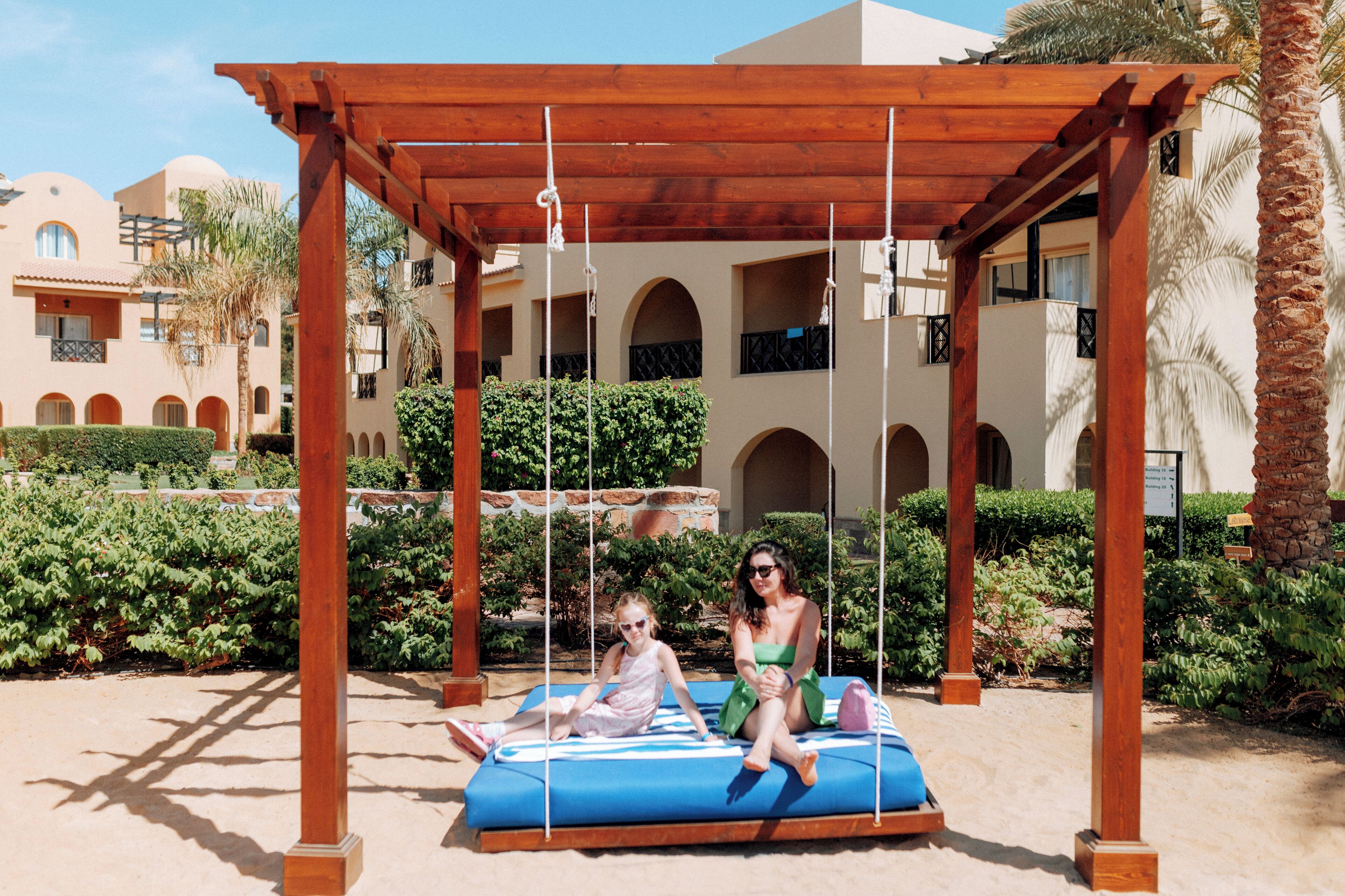 A wooden swing set with two people and a child, surrounded by greenery and a building with arches.