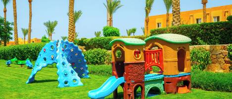 Un parque infantil con un tobogán azul y estructuras de escalada, rodeado de palmeras y un edificio al fondo.