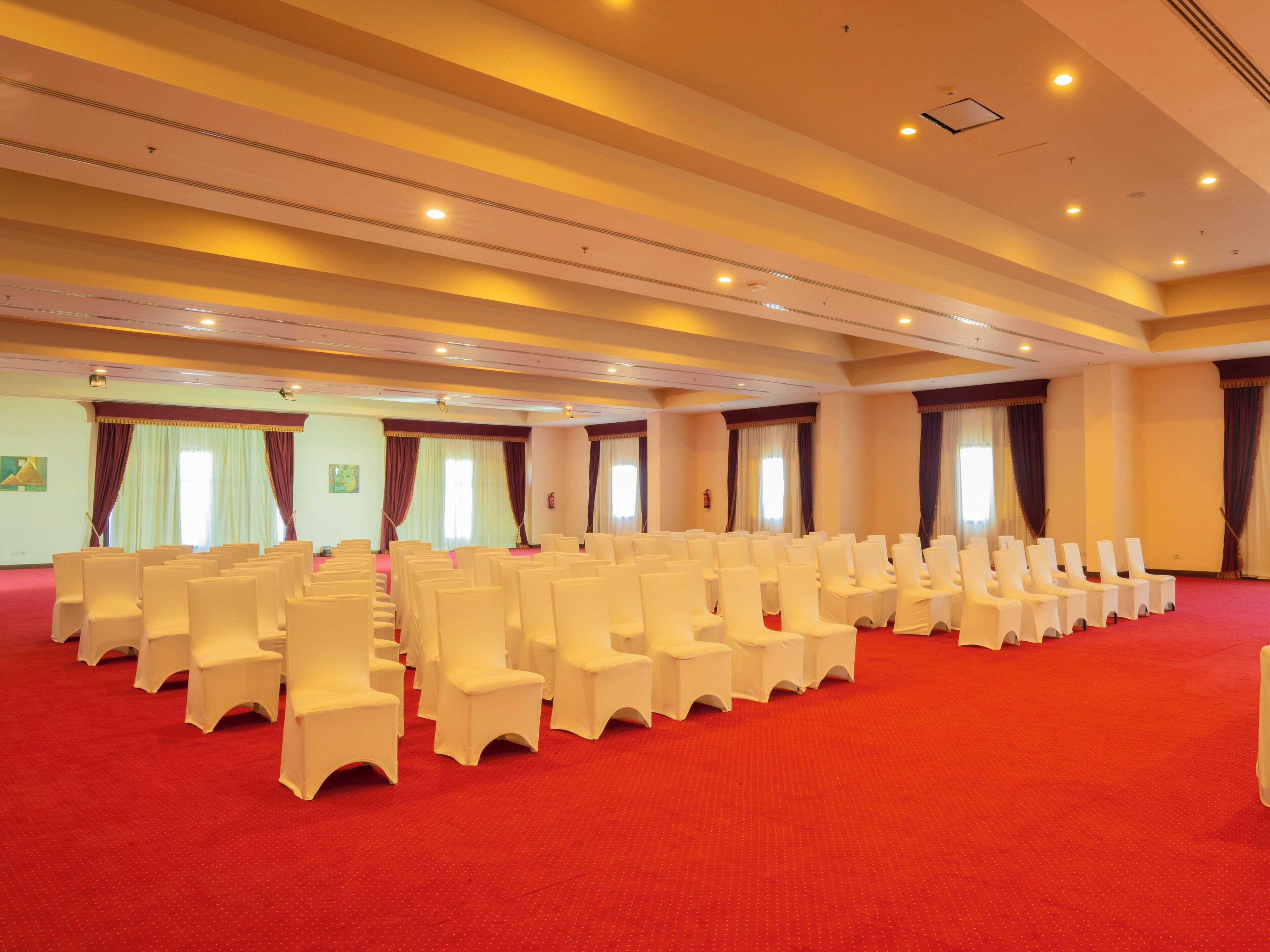 A conference room with rows of white chairs and a red carpet, large windows with curtains, and a high ceiling with recessed lighting.