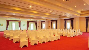 A conference room with rows of white chairs and a red carpet, large windows with curtains, and a high ceiling with recessed lighting.
