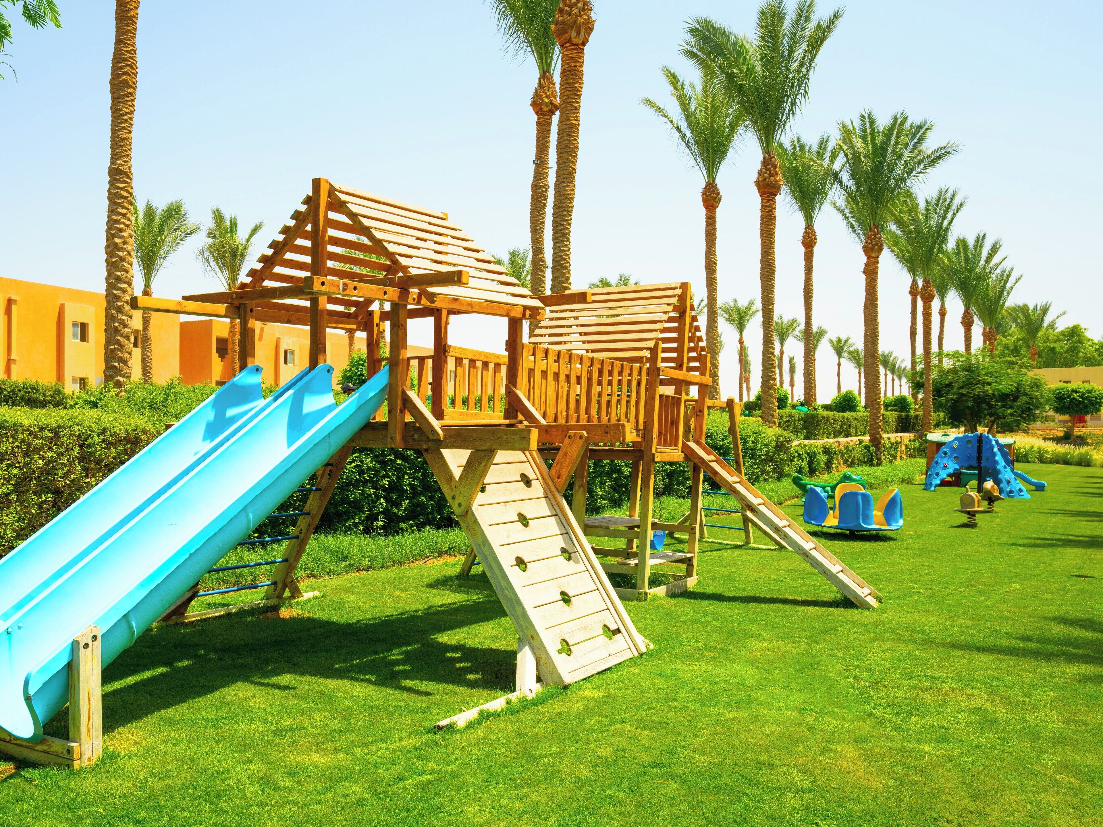 Un parque infantil con tobogán y estructuras para escalar, rodeado de palmeras y césped cuidado.