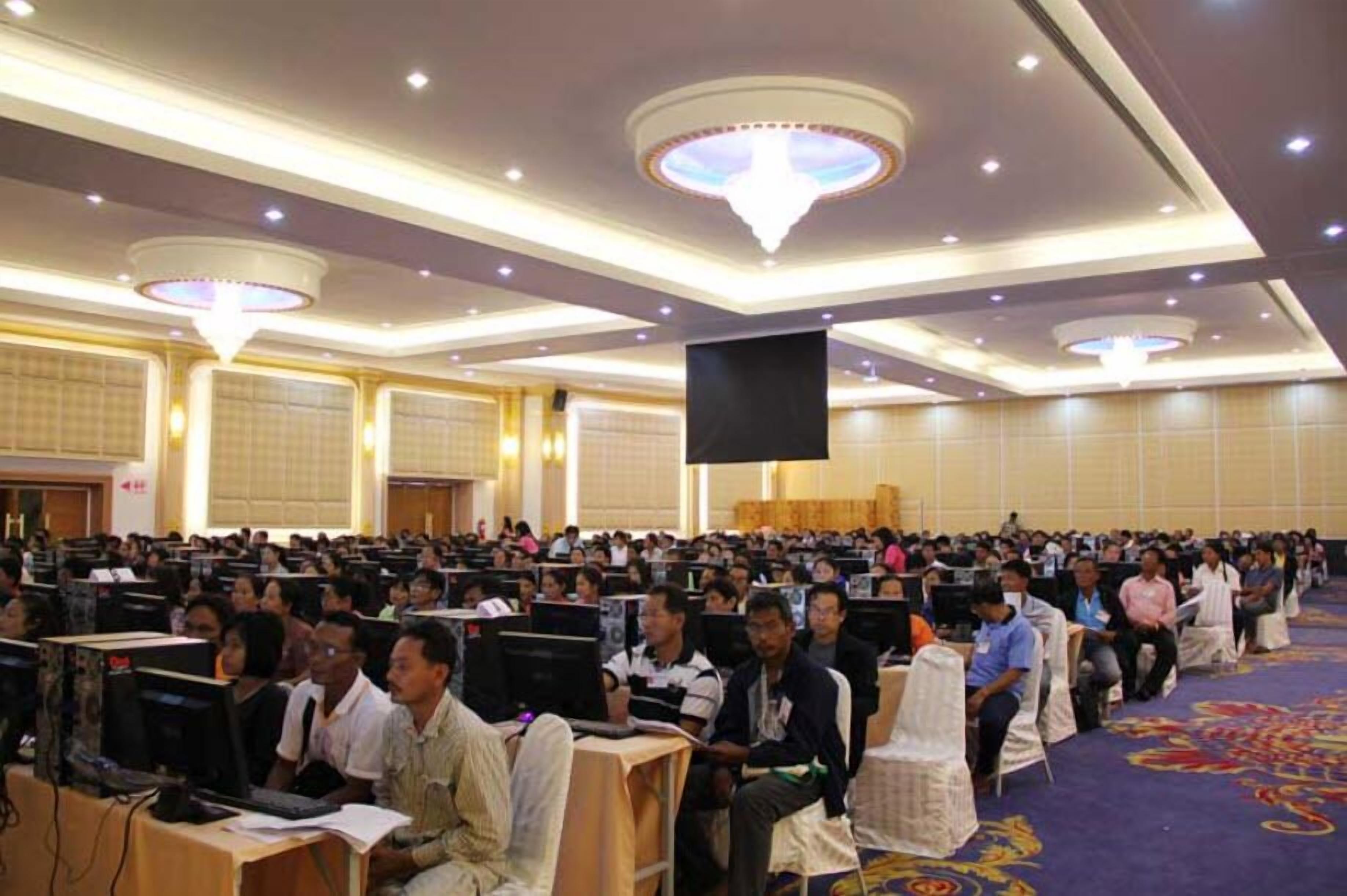 Une salle de conférence avec des rangées de tables et de chaises, un grand écran à l’avant et un lustre.