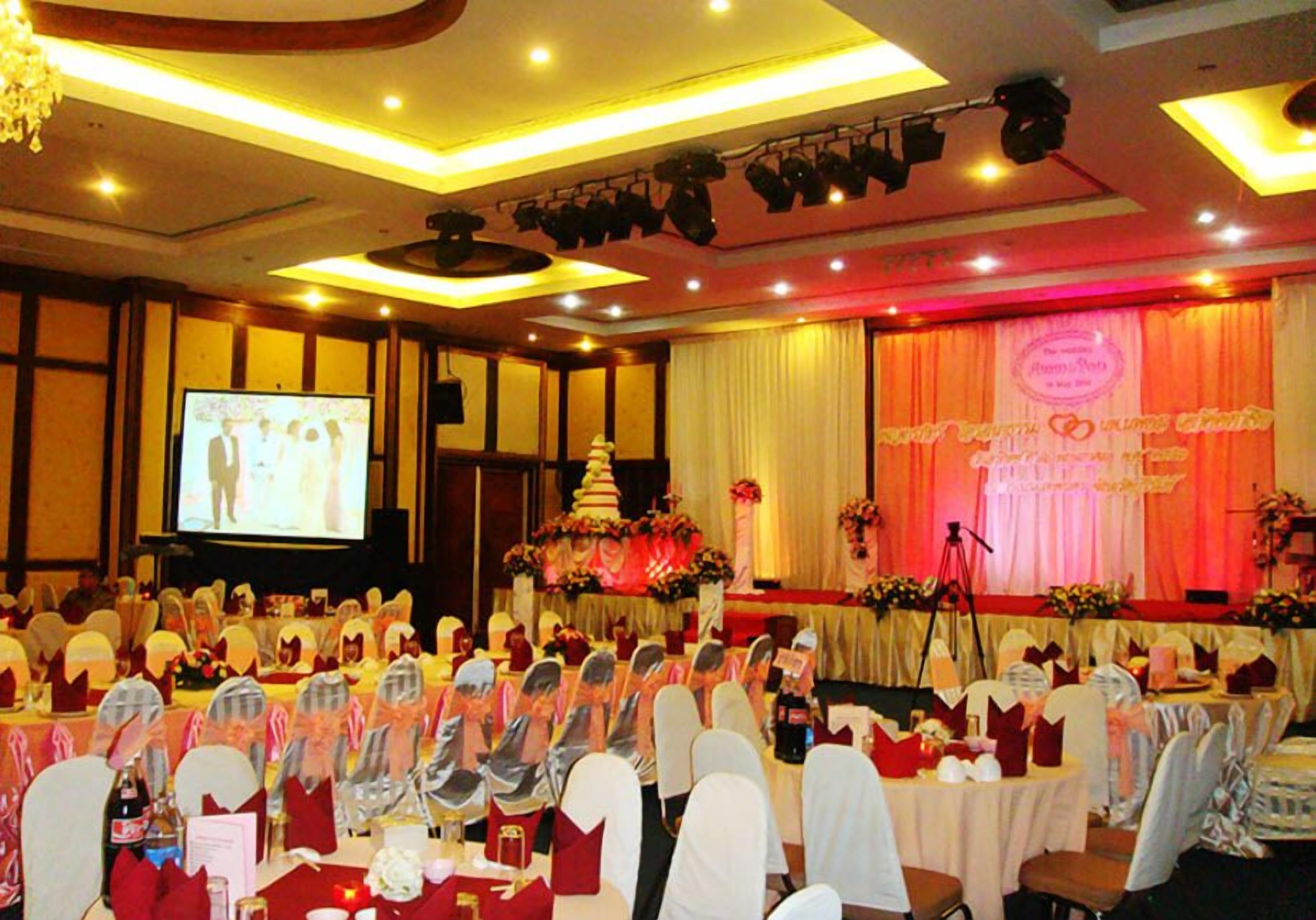 Un salón de banquetes con mesas redondas, sillas y un escenario decorado con un pastel y flores.