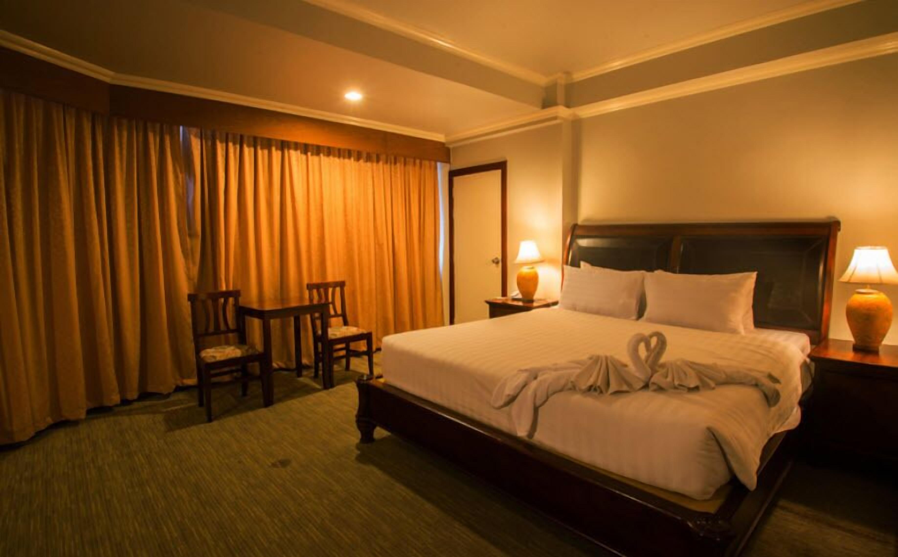 A hotel room with a large bed, two bedside lamps, a desk with two chairs, and a large window with curtains.