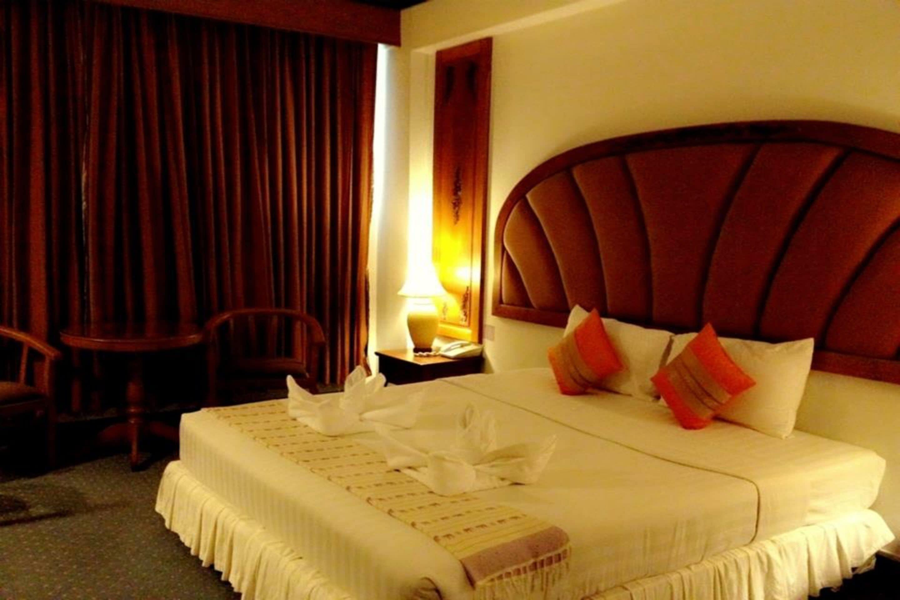 Una habitación de hotel con una cama grande, cabecera de madera, mesita de noche con lámpara y una zona de estar con sillas.