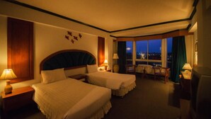 Habitación de hotel con dos camas, una mesita, sillas y vista a la ciudad de noche.