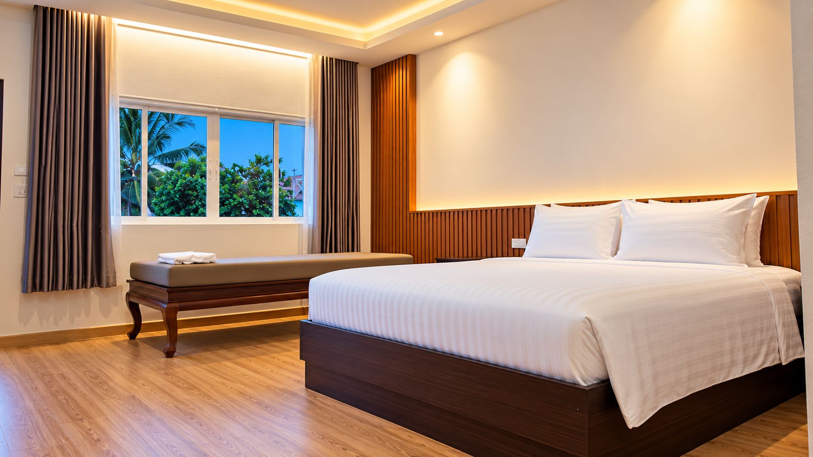 A hotel room with a large bed, a bench, a window with curtains, and a view of greenery outside.