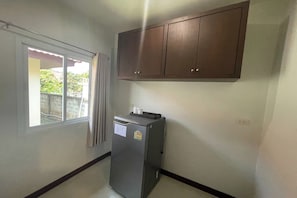 A compact kitchen with a refrigerator, a window with curtains, and a wooden cabinet above. - Phuket Airport Villa (Sa Khu)