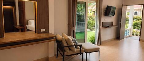 A modern room with a wooden desk, a cushioned chair, a television mounted on the wall, and a sliding glass door leading to a balcony.