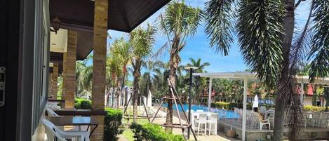 Un área de estar al aire libre techada, con sillas y mesas de plástico blanco, una piscina y palmeras.