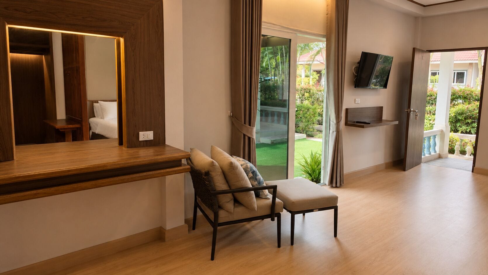 Une chambre d’hôtel moderne avec un bureau en bois, un fauteuil rembourré, un téléviseur à écran plat fixé au mur et une porte-fenêtre coulissante donnant sur un balcon.