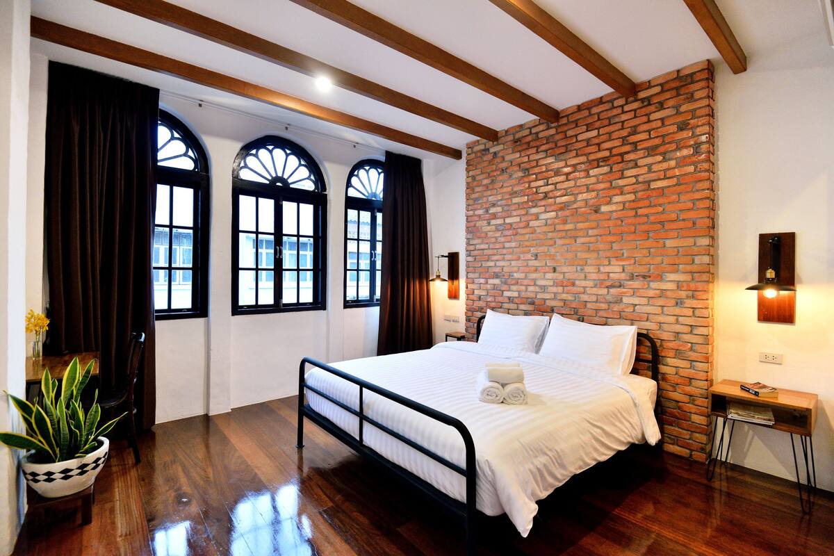 A bedroom with a brick wall, a bed with white linens, a nightstand, a potted plant, and a window with curtains.