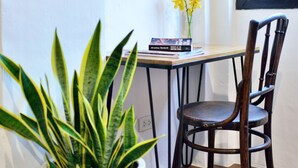 Una habitación con piso de madera, una planta con estampado blanco y negro en una maceta blanca, una silla de madera, una mesita con libros y un jarrón con flores amarillas, y una ventana con vista al exterior.