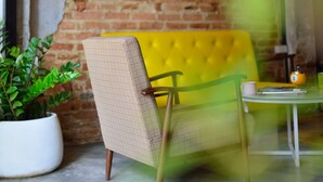 Una habitación con pared de ladrillo, sofá amarillo, sillón de mimbre, una mesita y una planta en maceta.