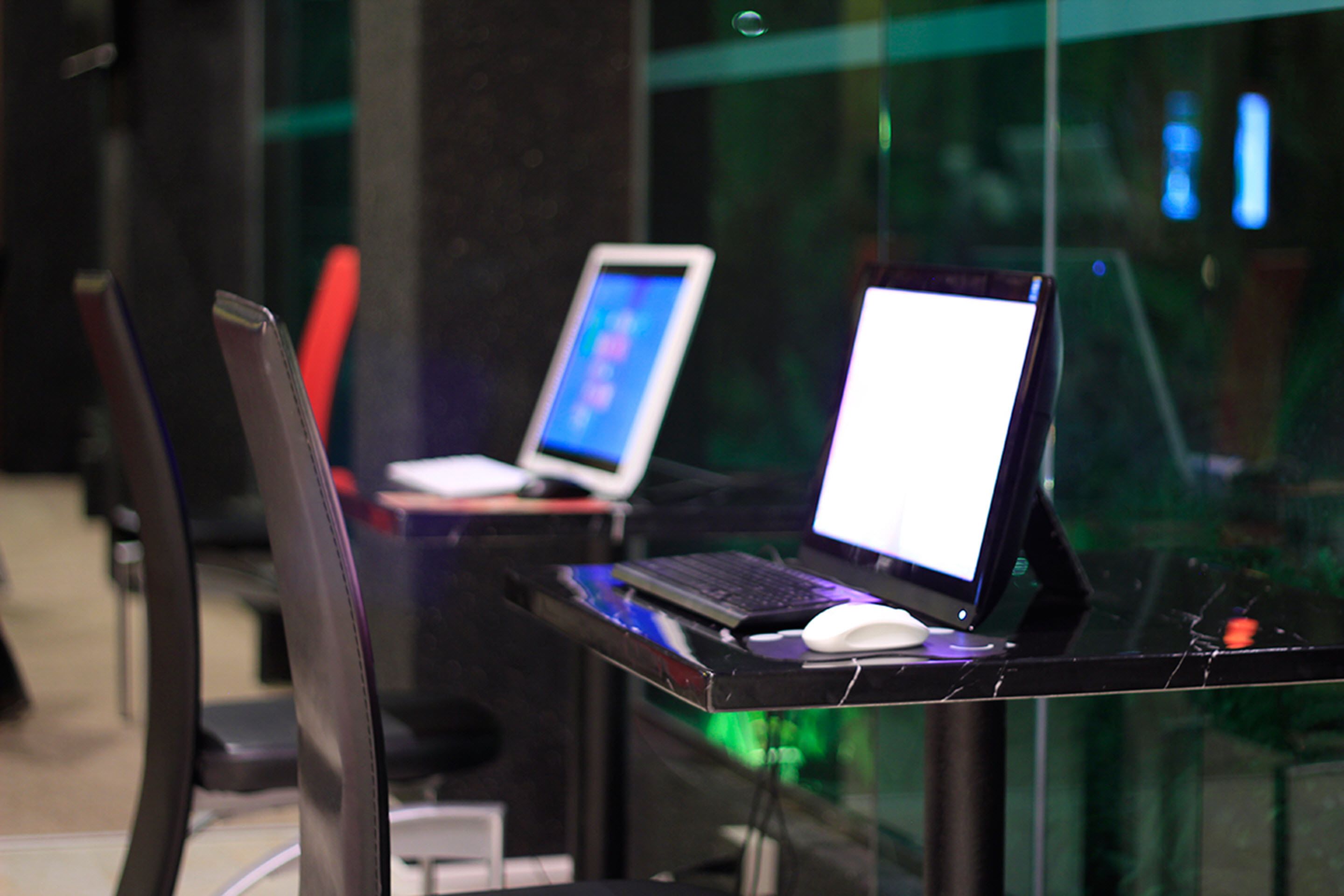 A modern workspace with two laptops on a glass table, a white mouse, and a black chair.