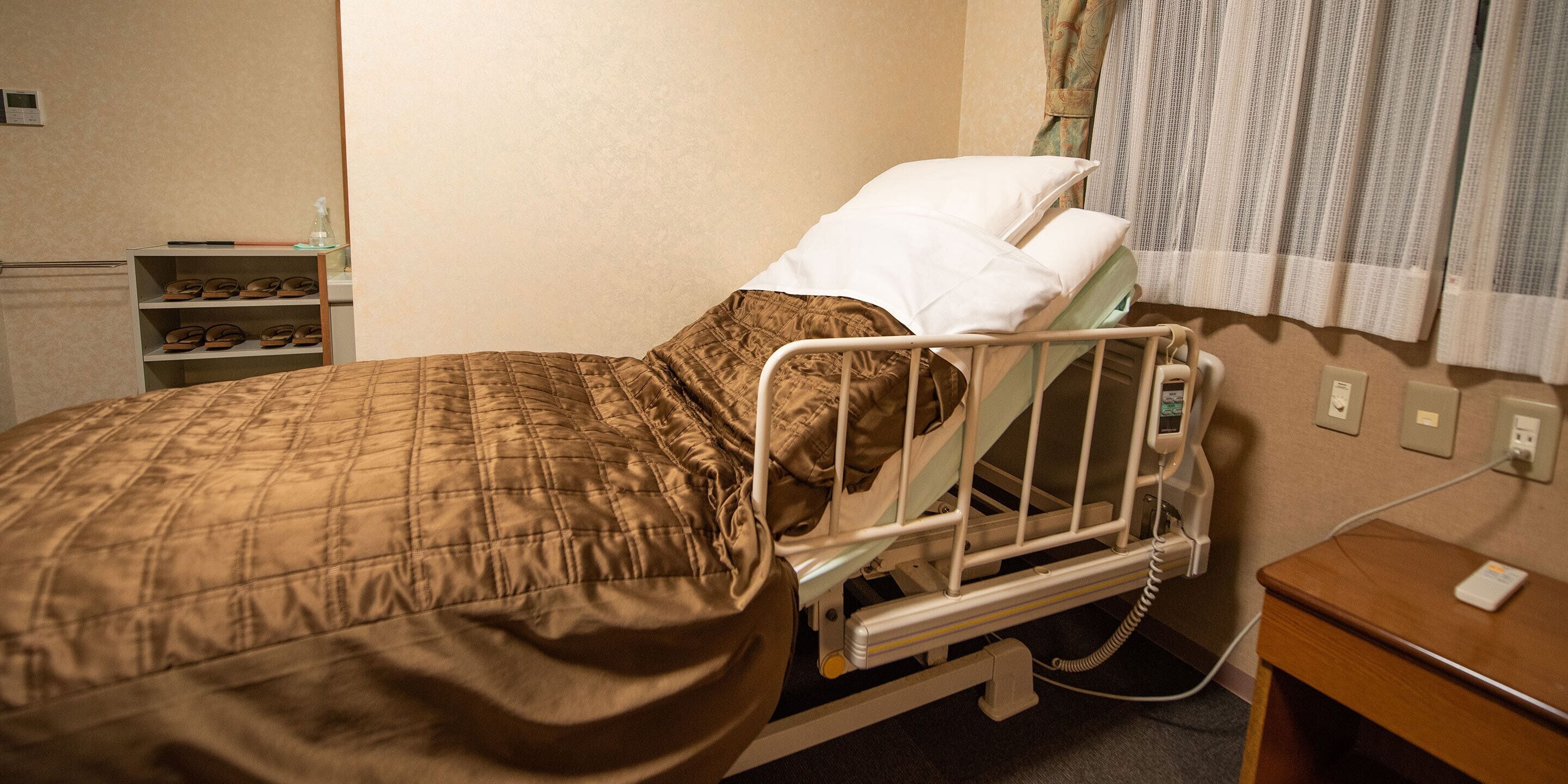 Una habitación de hospital con una cama, almohadas, una mesita de noche y un estante con zapatos.
