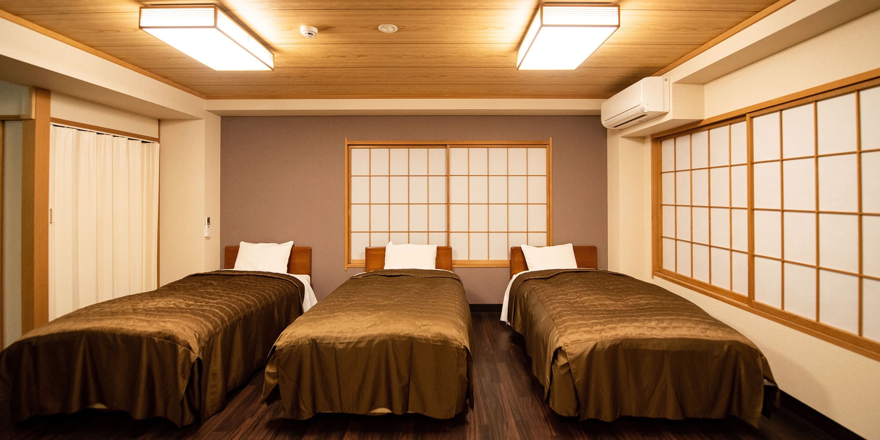 Une chambre d’hôtel avec trois lits simples, un plancher en bois, un paravent de style japonais et un plafond doté d’un éclairage encastré.