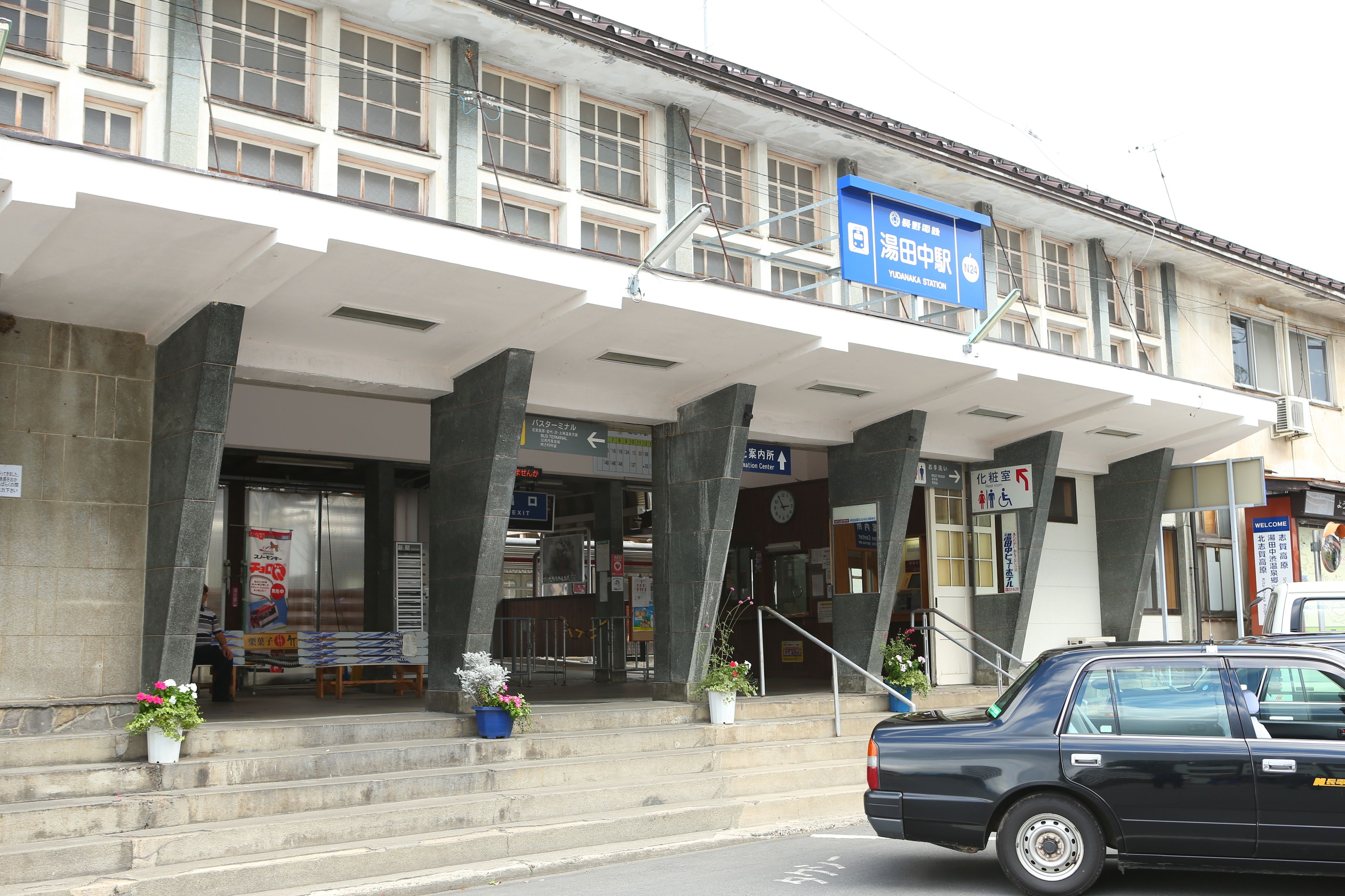 A building with a blue sign indicating a station entrance, a staircase leading to the entrance, and a parked car in front.