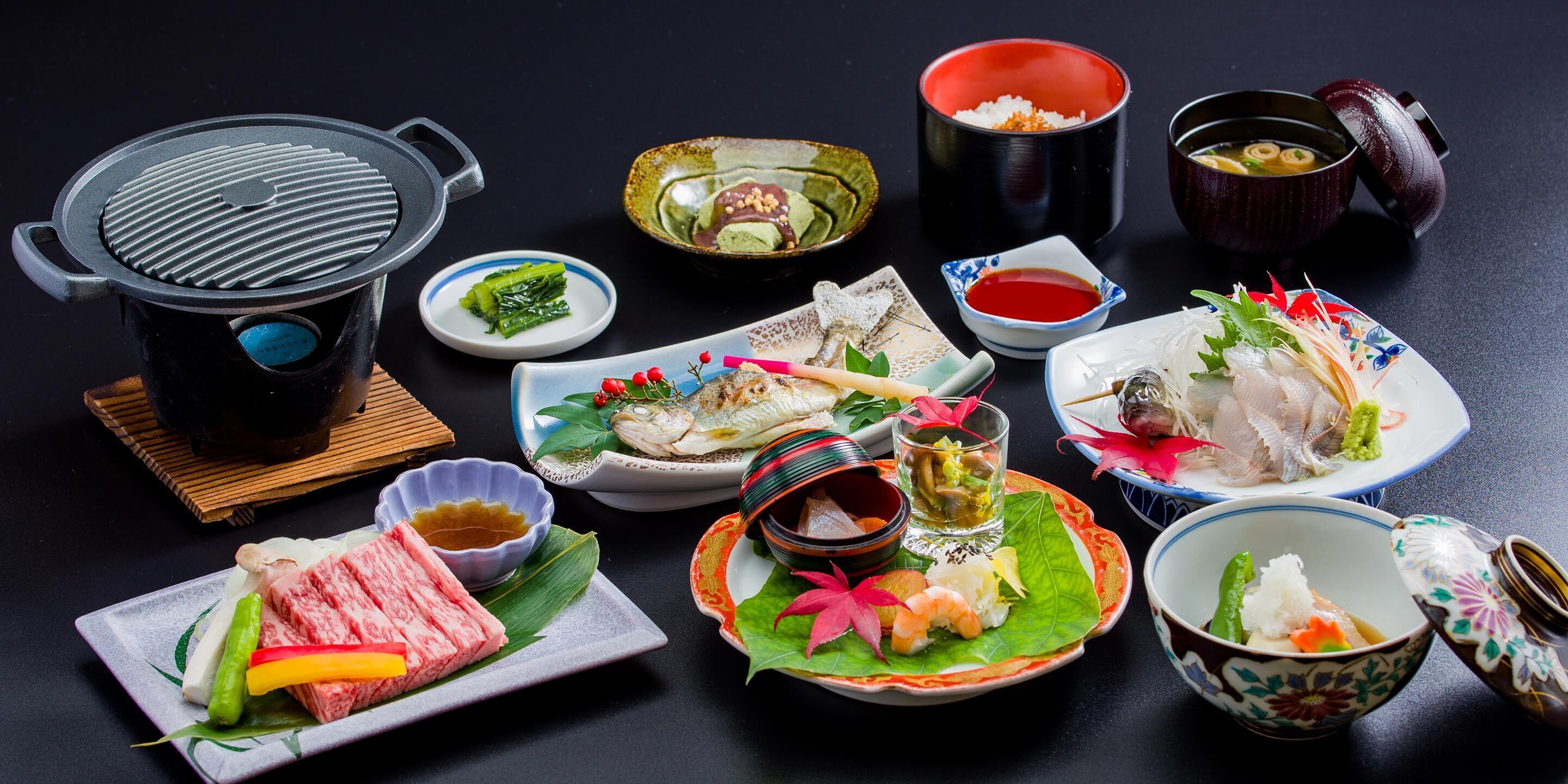 A variety of Japanese dishes including sushi, sashimi, and a hot pot, arranged on a dark surface.