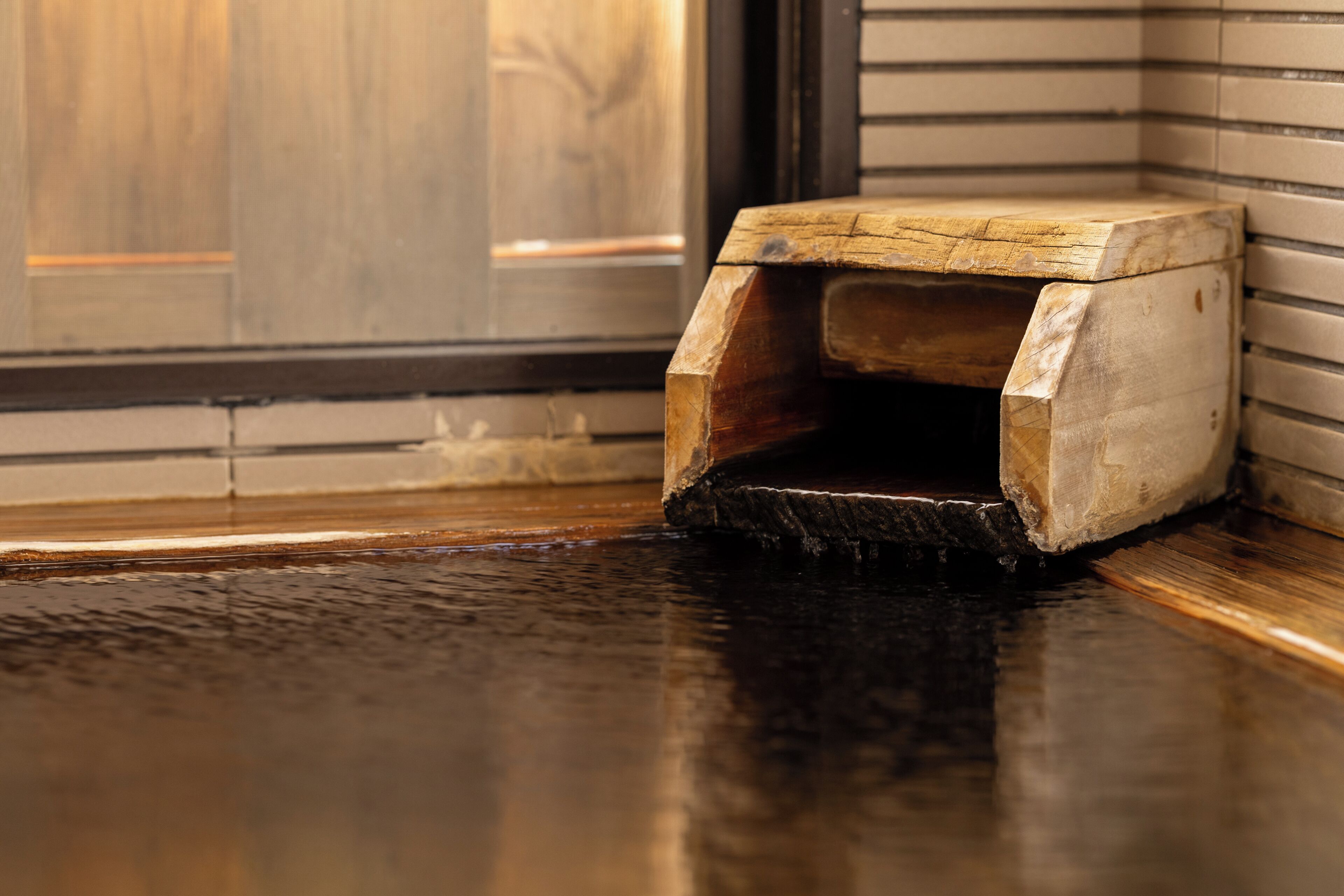 A wooden corner bench with a slatted design, positioned against a wall with a tiled floor.