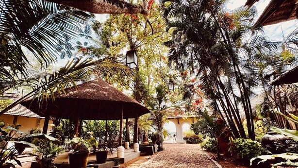 Un chemin mène à un bâtiment dont le toit est recouvert de chaume, et qui est entouré d’une végétation luxuriante et de plantes en pot.