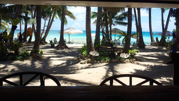 A beach view with palm trees, a sign reading "DEPARTIS RESORT", and a sandy area with chairs and umbrellas. - Deparis Beach Resort (Boracay Island)