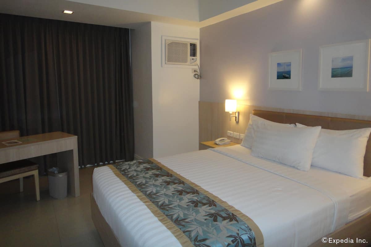 A hotel room with a bed, a desk, a chair, a waste bin, a nightstand with a lamp, and two framed pictures on the wall.