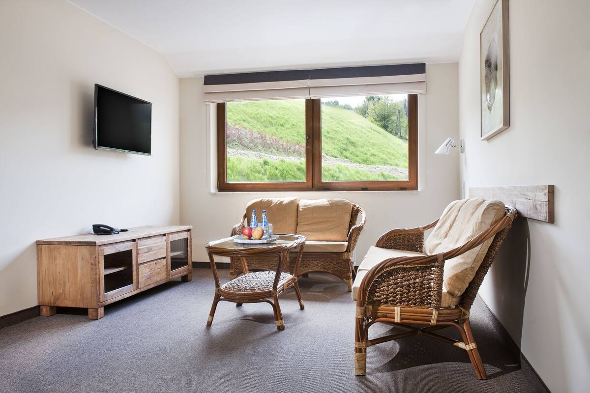 Ein Zimmer mit Flachbildfernseher, Korbmöbeln, einem kleinen Tisch mit Obst und Flaschen sowie Blick auf eine grüne Hügellandschaft durch ein großes Fenster.