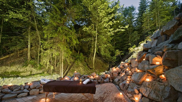 Une terrasse en bois avec un banc, entourée de rochers et d’arbres, éclairée par des bougies qui créent une ambiance chaleureuse.