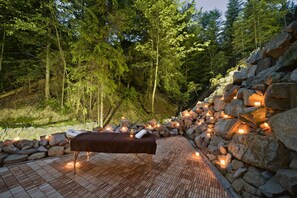 A wooden deck with a bench, surrounded by rocks and trees, with candles providing ambient lighting.