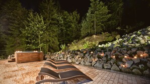 Une terrasse extérieure dotée de chaises longues et d’un jacuzzi, entourée d’un mur de pierre et d’arbres.