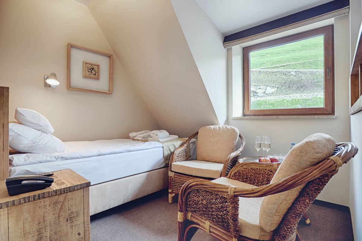 Ein Schlafzimmer mit einem Bett, zwei Korbstühlen, einem Nachttisch, einem gerahmten Bild und einem Fenster mit Blick auf eine Wiese.