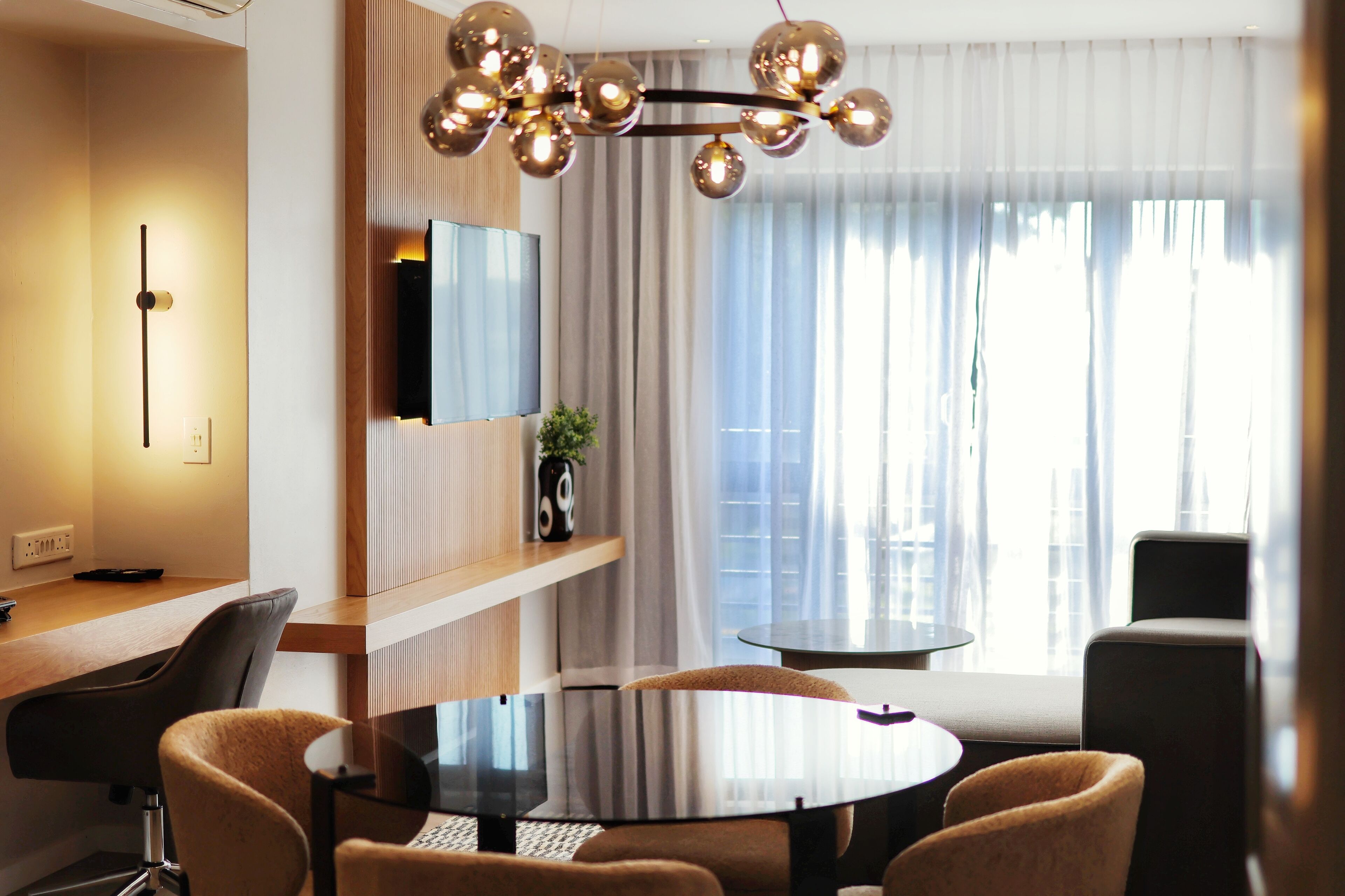 Una habitación de hotel moderna con una mesa redonda para comer, asientos cómodos, un televisor montado y un amplio ventanal con cortinas.