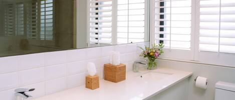 A bathroom with white tiled walls, a white countertop, a mirror, a sink, a toilet, and a window with blinds.