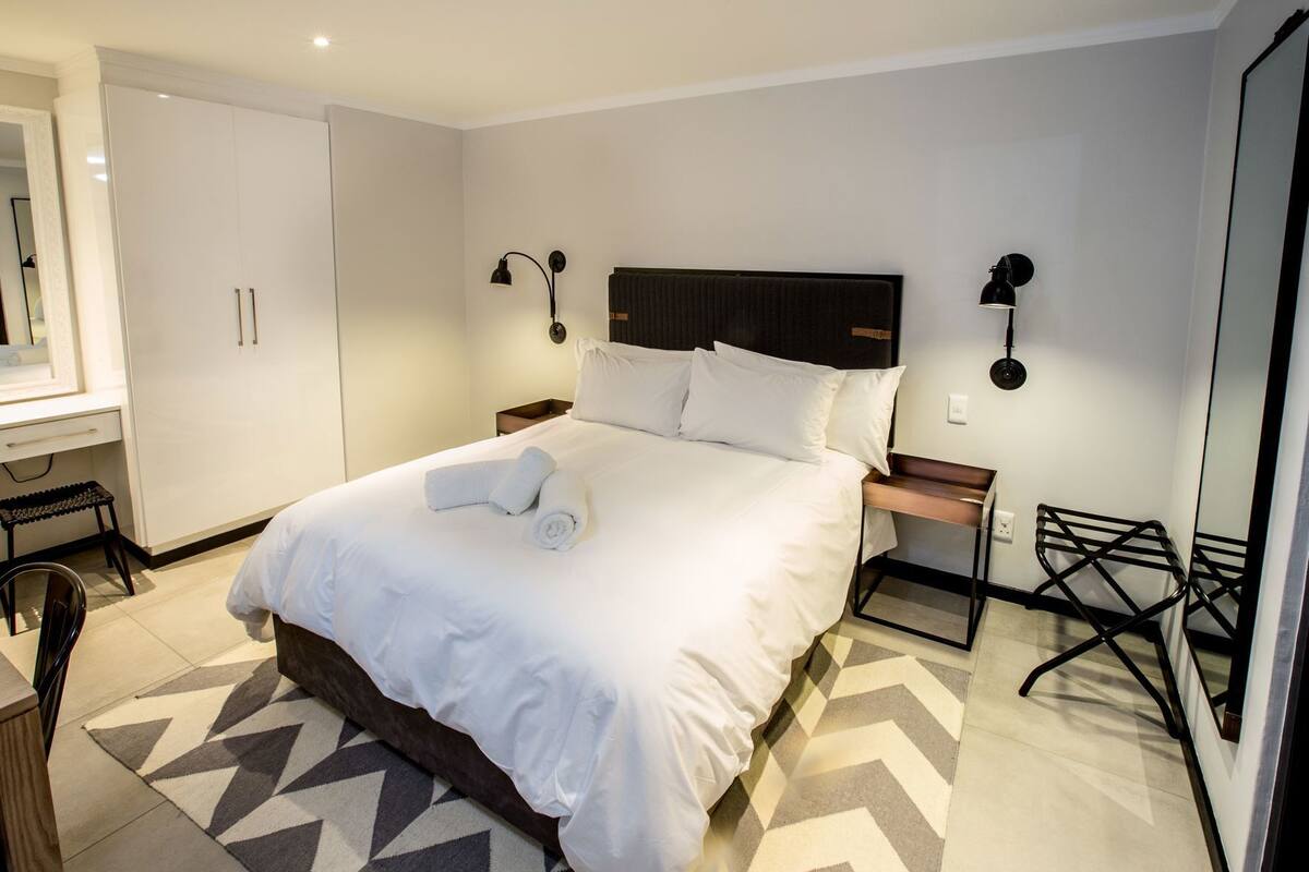 A neatly made bed with white linens, a headboard, bedside tables, a chair, and a mirror.