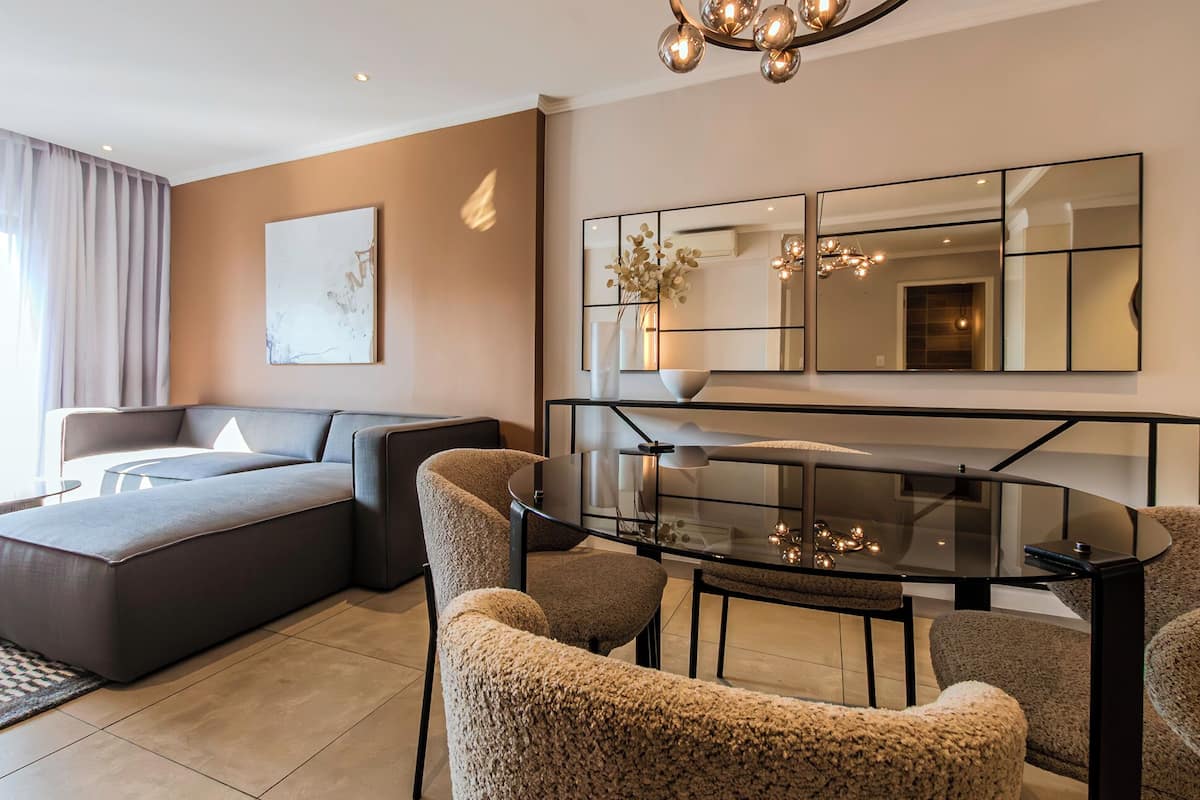 A modern living room with a sofa, ottoman, and a glass dining table with chairs. There is a large mirror and artwork on the wall.