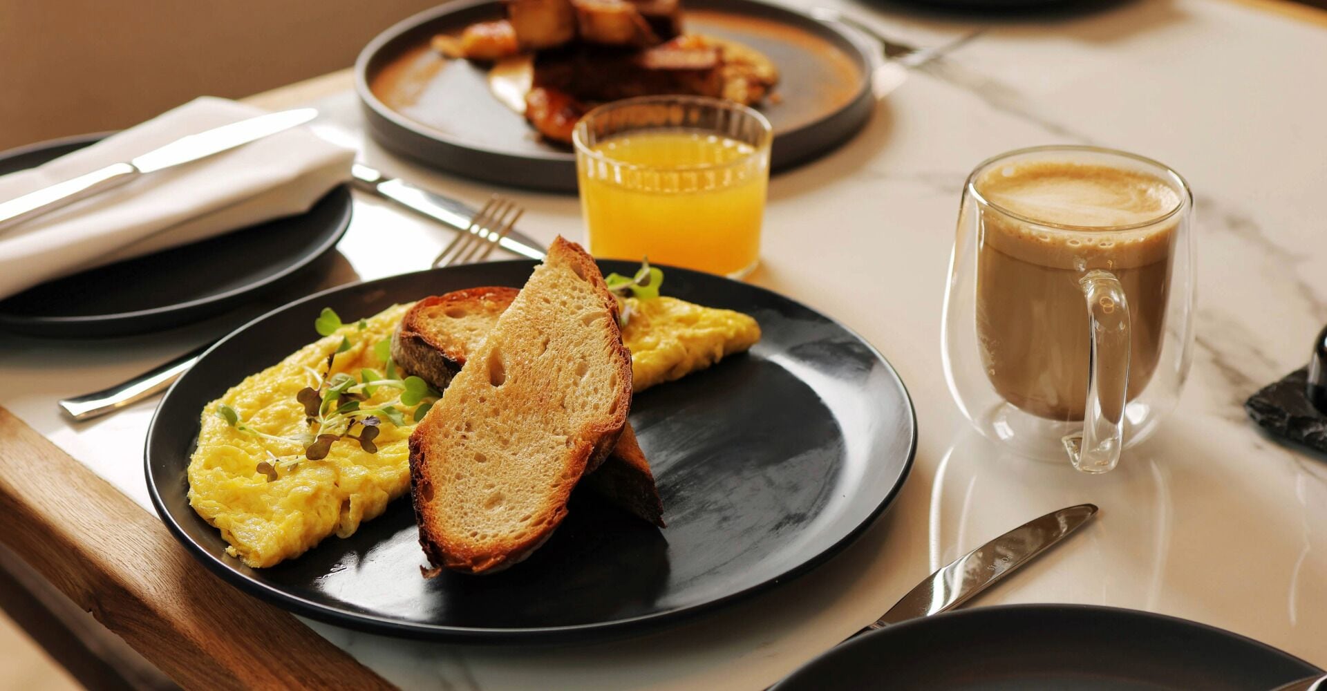 Une table en marbre dressée pour le petit-déjeuner, avec des œufs brouillés, des toasts, du jus d’orange et du café.
