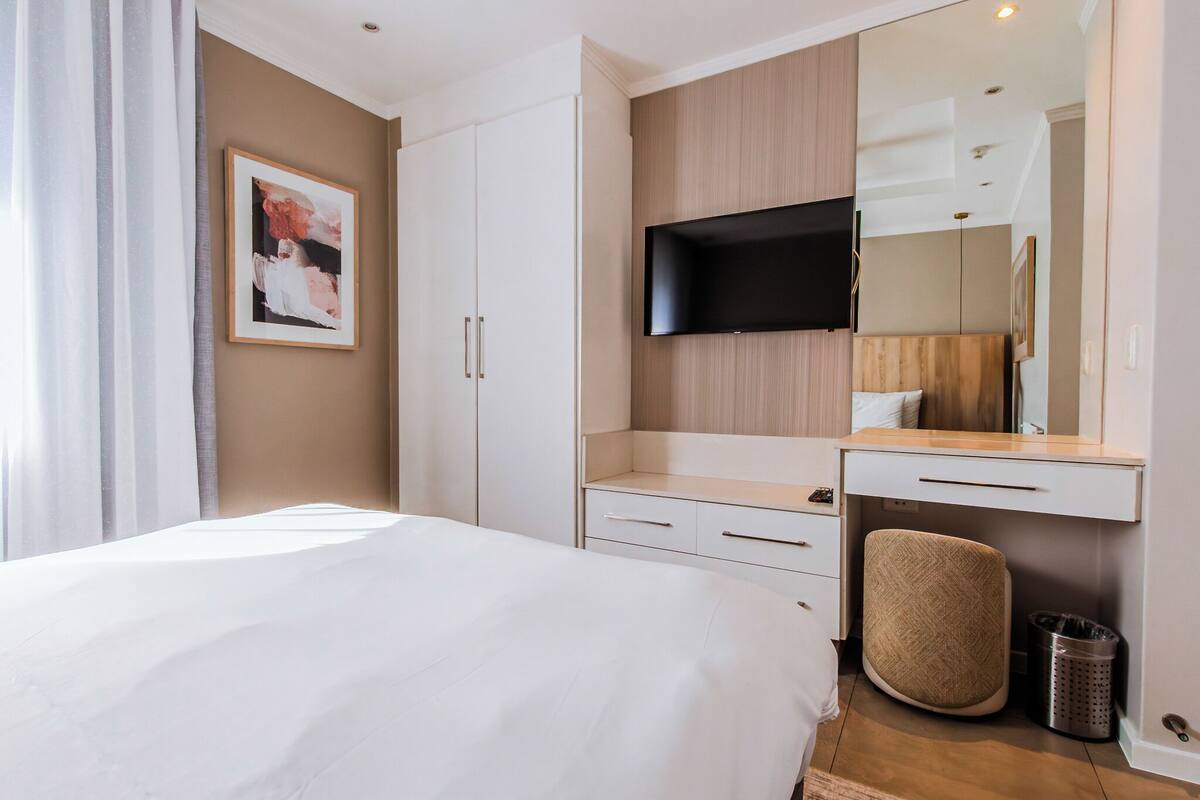 A modern hotel room with a large bed, a flat-screen TV mounted on the wall, a wooden vanity with a mirror, and a small trash bin.