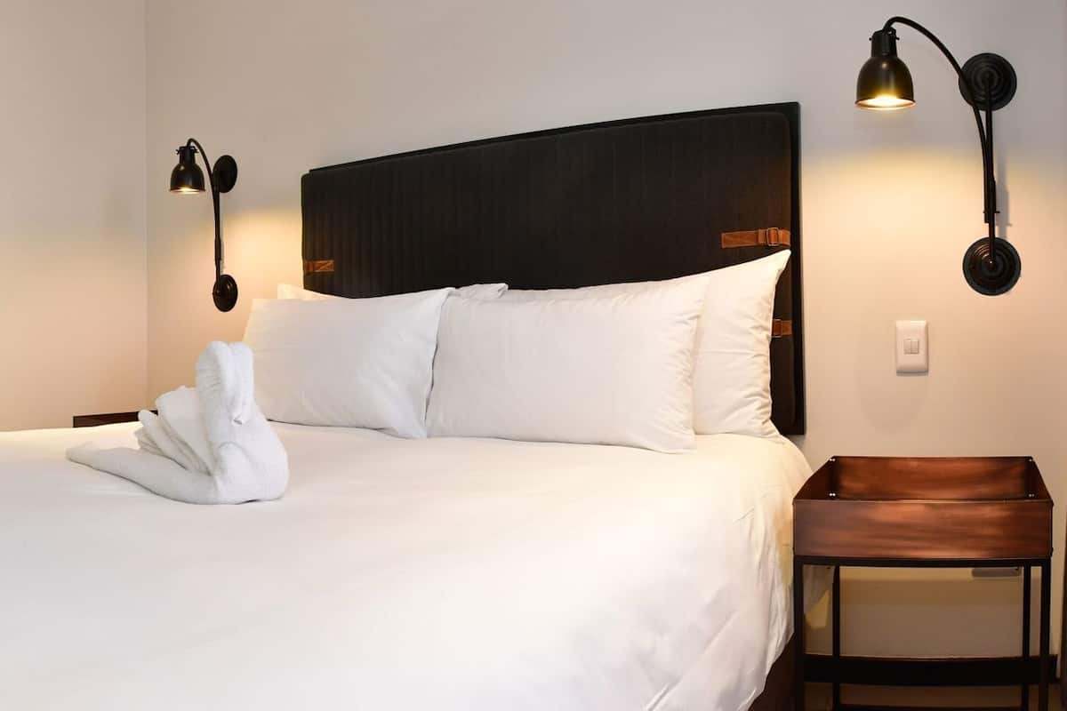 A hotel room with a large bed, a desk with drawers, a trash bin, and a nightstand with a lamp.