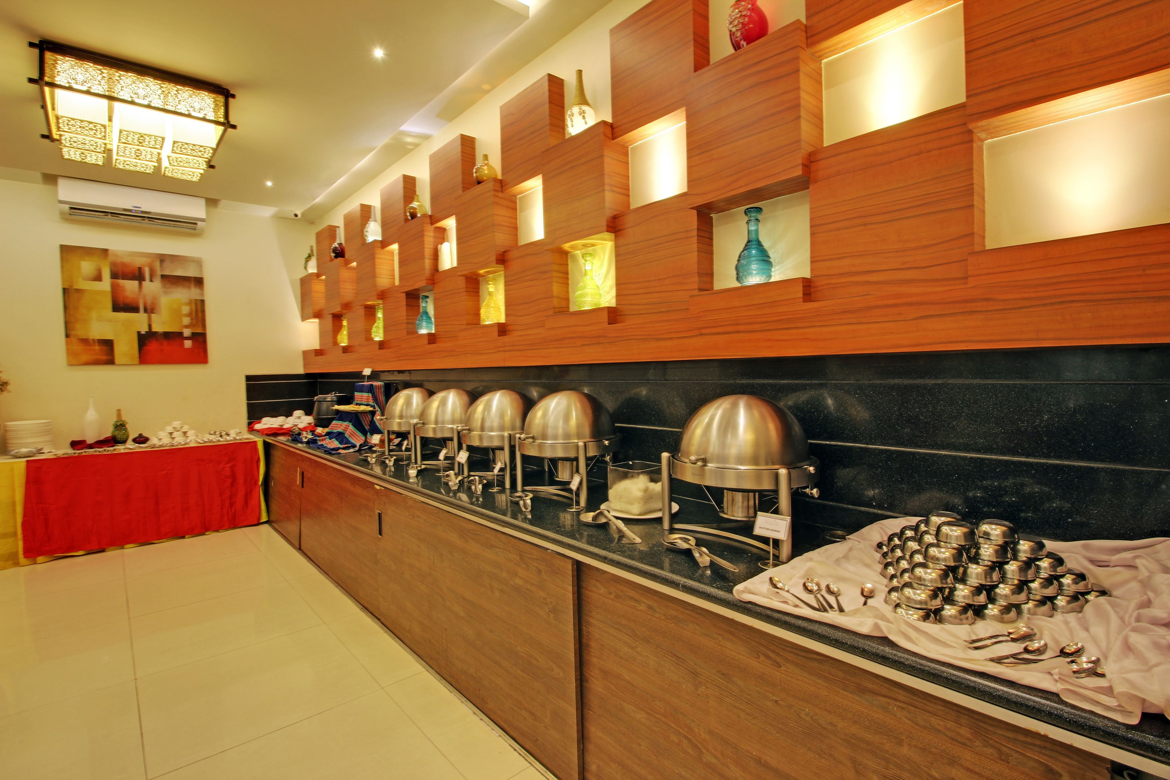 A buffet setup with multiple serving stations, a red table with dishes, and a wall with shelves displaying bottles and a decorative piece.