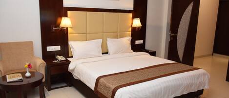 Una habitación de hotel con una cama, mesitas de noche, una silla, un cuadro en la pared y una puerta.