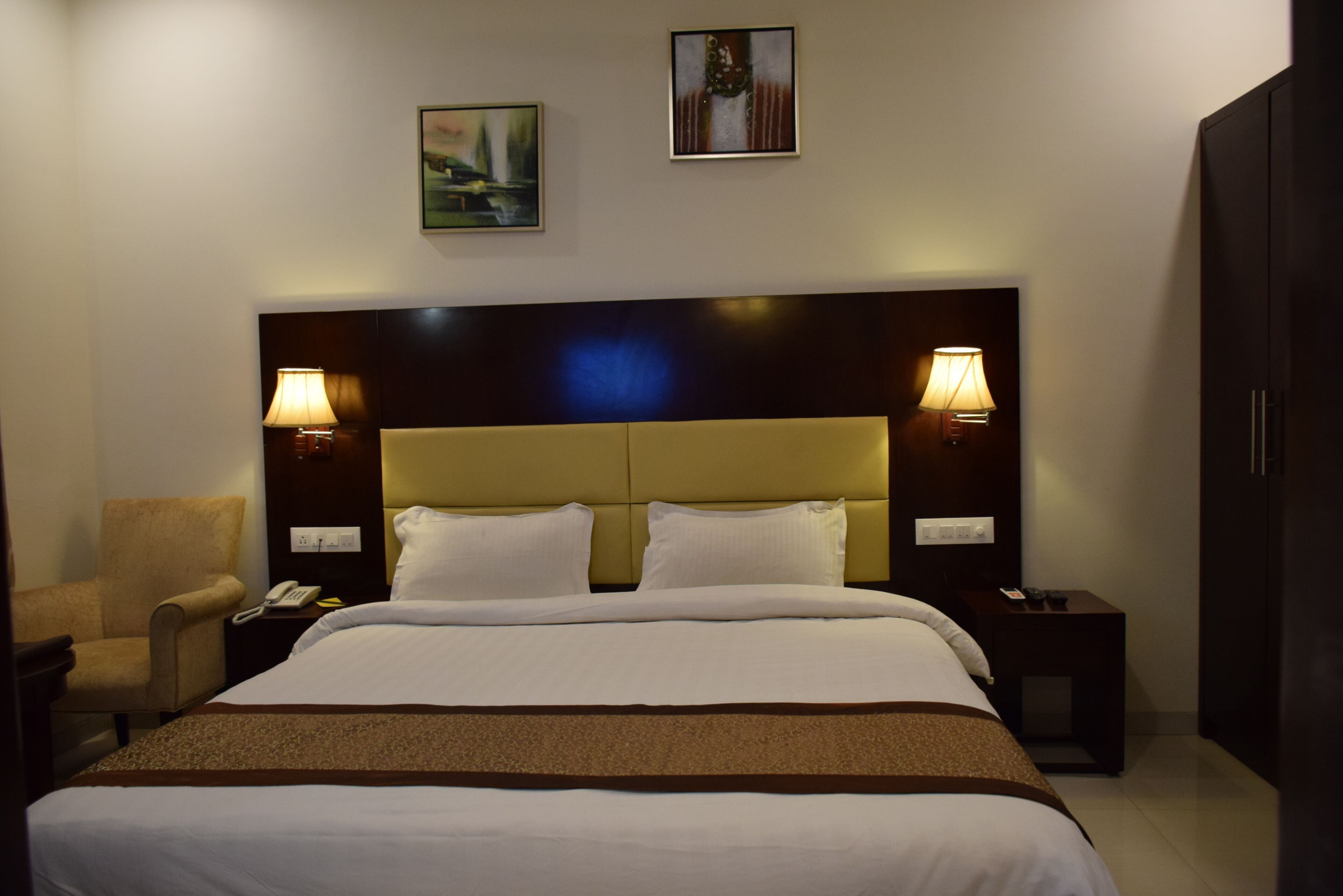 Een hotelkamer met een groot bed, twee bedlampen, een stoel, een nachtkastje met een telefoon, en twee foto's in lijsten aan de muur.