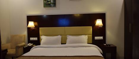 Een hotelkamer met een groot bed, twee bedlampen, een stoel, een nachtkastje met een telefoon, en twee foto's in lijsten aan de muur.